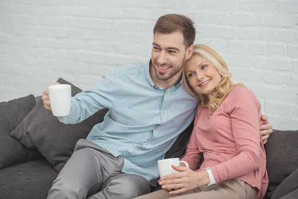 Porträt einer lächelnden Mutter und ihres erwachsenen Sohnes mit Tassen Heißgetränken zu Hause — Stockfoto