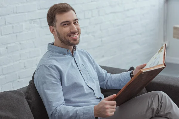 Retrato del hombre sonriente con álbum de fotos descansando en el sofá en casa - foto de stock