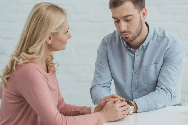 Madre e hijo adulto tomados de la mano mientras están sentados en la mesa - foto de stock