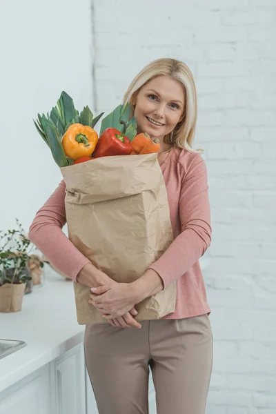 Portrait of smiling mature woman with paper bag full of fresh vegetables in hands at home — Stock Photo