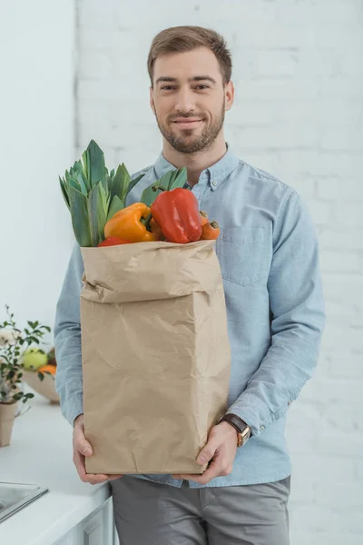 Retrato de hombre con bolsa de papel llena de verduras frescas para la cena en las manos en casa - foto de stock