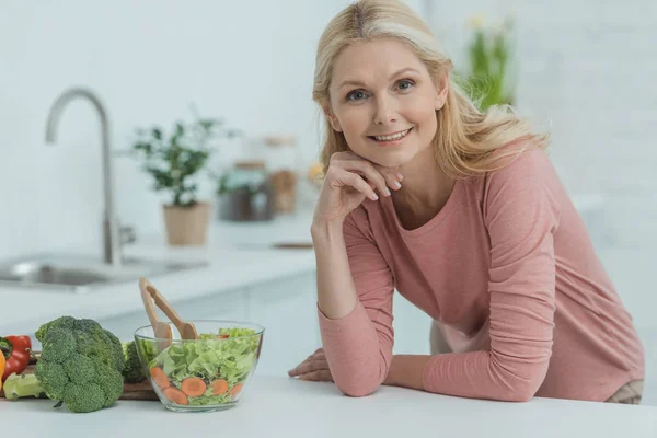 Retrato de mujer madura sonriente con ensalada fresca en el mostrador en la cocina - foto de stock