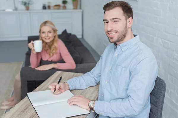 Selective focus of smiling man at table with notebook and mother with cup of coffee at home — Stock Photo