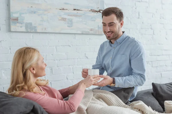 Side view of smiling careful son brought cup of tea to mother on sofa at home — Stock Photo
