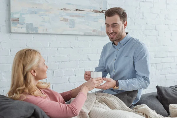 Vista laterale di sorridente figlio attento portato tazza di tè alla madre sul divano a casa — Foto stock