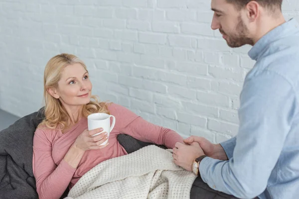 Mother with cup of tea lying on sofa with grown son near by at home — Stock Photo