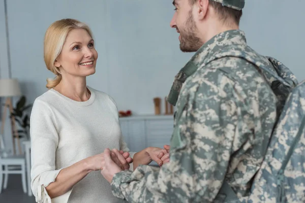 Mère souriante et fils adulte en uniforme militaire tenant la main ensemble à la maison — Photo de stock