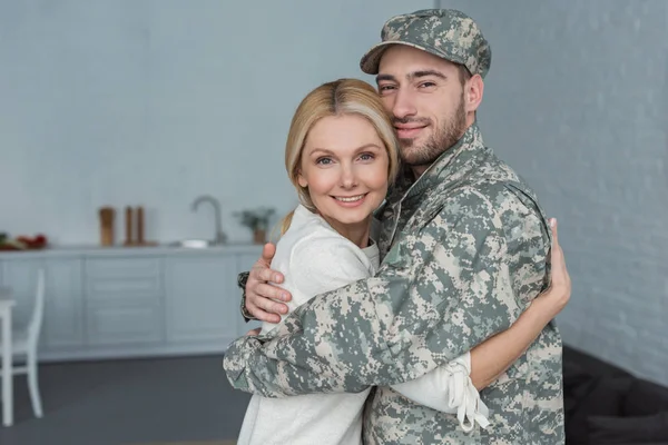 Retrato del hombre sonriente en uniforme militar y la madre abrazándose en casa - foto de stock