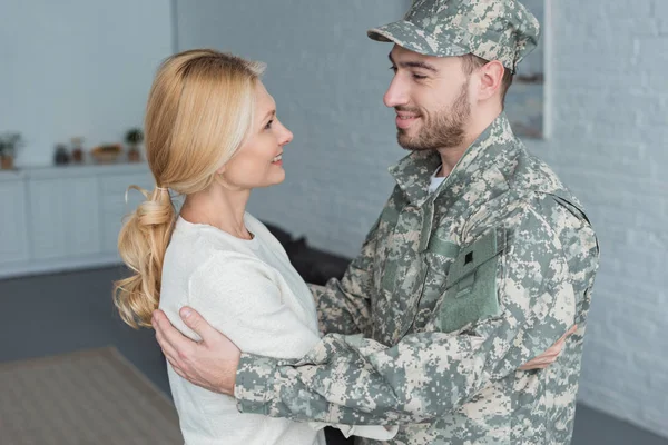 Vista lateral de homem sorridente em uniforme militar e mãe abraçando uns aos outros em casa — Fotografia de Stock