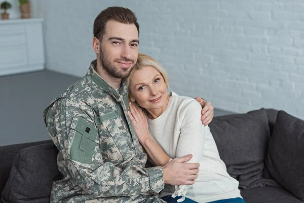 Retrato del hombre en uniforme militar abrazando sonriente madre en el sofá en casa - foto de stock