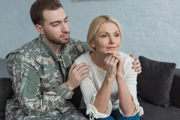 Retrato crescido filho no militar uniforme abraçando mãe pensativa no sofá em casa — Fotografia de Stock