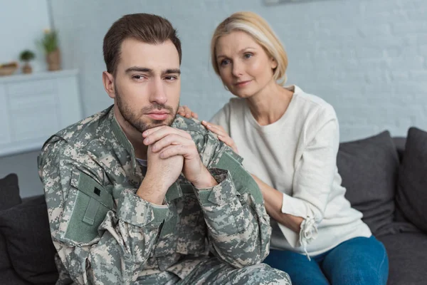 Retrato del hombre reflexivo en uniforme militar y la madre cerca en el sofá en casa - foto de stock