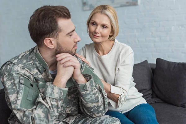 Retrato del hombre reflexivo en uniforme militar y la madre cerca en el sofá en casa - foto de stock