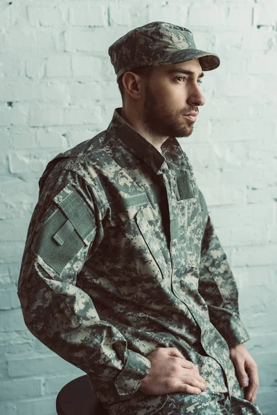 Porträt eines nachdenklichen Soldaten in Militäruniform, der auf einem Stuhl vor einer weißen Backsteinwand sitzt — Stockfoto