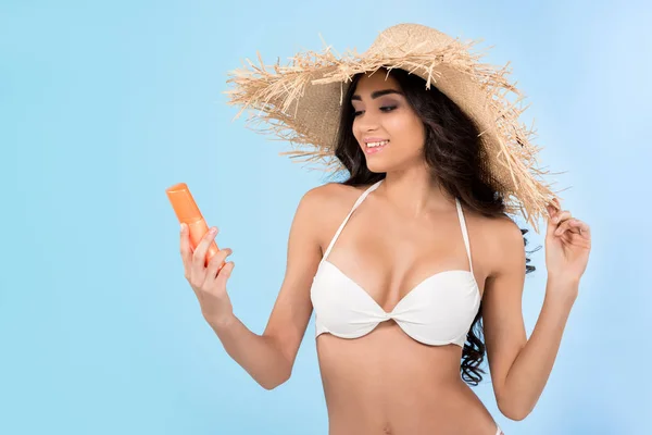 Ragazza sorridente in bikini guardando una bottiglia di crema solare, isolata sul blu — Foto stock