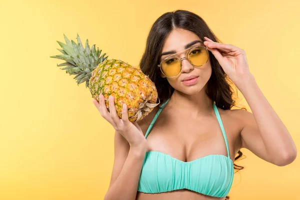 Attractive girl in sunglasses posing with fresh pineapple, isolated on yellow — Stock Photo