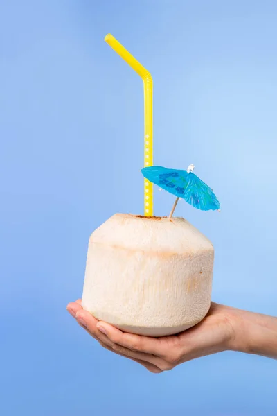 Cropped view of female hand with summer cocktail in fresh coconut with umbrella and straw, isolated on blue — Stock Photo