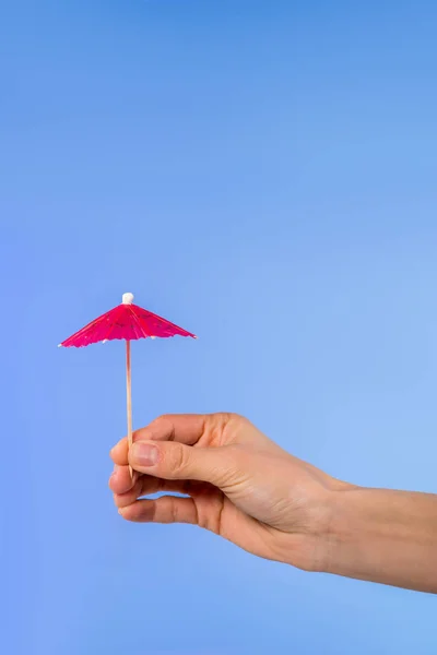 Cropped view of female hand with cocktail umbrella, isolated on blue — Stock Photo