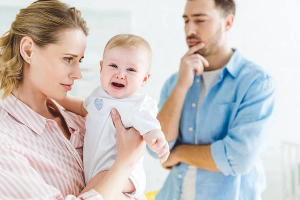 Mother holding crying infant daughter on hands and thoughtful father standing near — Stock Photo