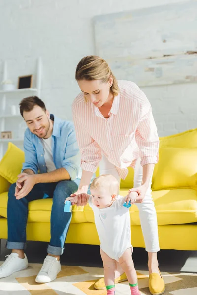 Madre enseñando a la hija bebé a caminar y el padre sentado en el sofá en la habitación moderna - foto de stock