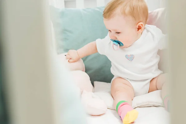 Infant with baby dummy and teddy hare sitting in crib — Stock Photo