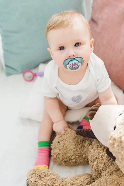 High angle view of infant with baby dummy and teddy bear sitting in crib — Stock Photo