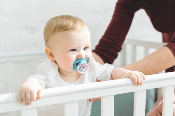 Cropped image of mother holding infant daughter with baby dummy standing in crib — Stock Photo