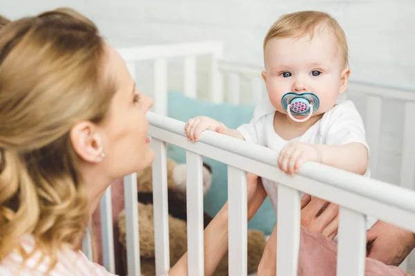 Mother holding infant daughter with baby dummy standing in crib — Stock Photo