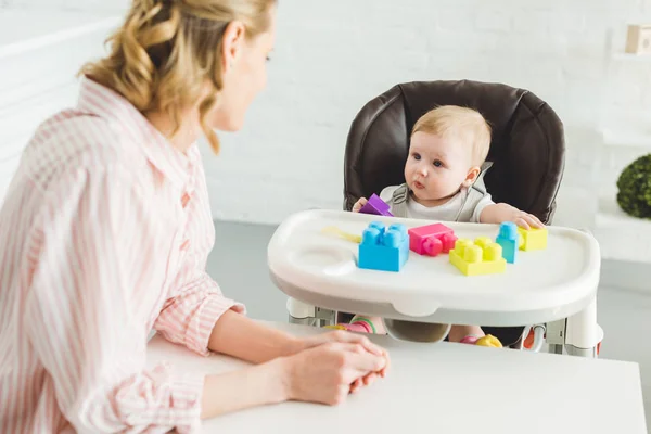 Mãe assistindo na filha infantil sentada em cadeira de bebê com blocos de plástico — Fotografia de Stock