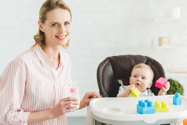 Young mother with feeding bottle and infant daughter sitting in baby chair with plastic blocks — Stock Photo