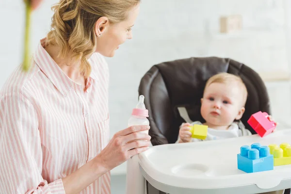 Young mother holding feeding bottle and infant daughter sitting in baby chair with plastic blocks — Stock Photo