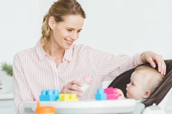 Mother with feeding bottle and infant daughter sitting in baby chair with plastic blocks — Stock Photo