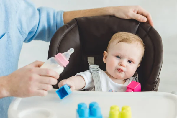 Обрезанный вид отца, держащего кормящую бутылочку и младенца дочь, сидящую в детском кресле с пластиковыми блоками — стоковое фото