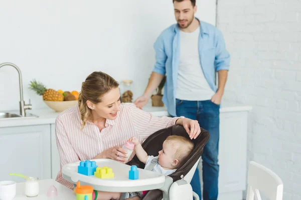 Mutter gibt Säuglingstochter im Kinderstuhl Fütterungsflasche, während Vater dahinter steht — Stockfoto