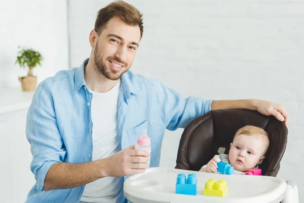 Smiling father with feeding bottle and infant daughter in baby chair with plastic blocks — Stock Photo