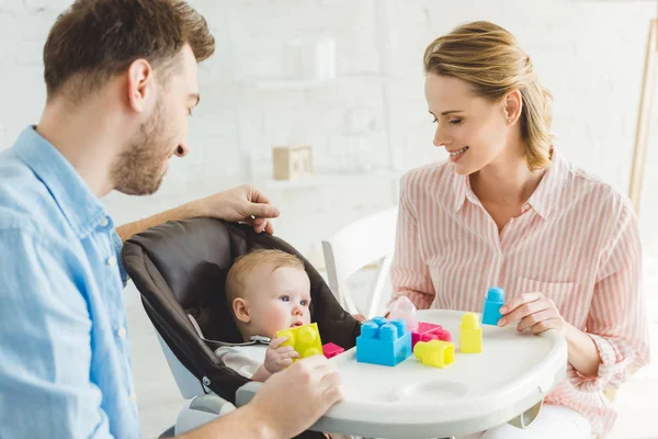 Smiling parents and infant daughter in baby chair with plastic blocks — Stock Photo