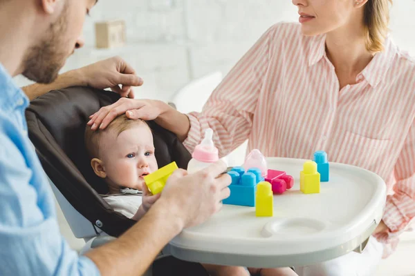 Cropped view of parents with feeding bottle and infant daughter in baby chair with plastic blocks — Stock Photo