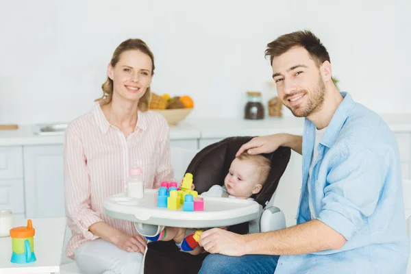 Young parents with infant daughter sitting in baby chair with plastic blocks and feeding bottle — Stock Photo