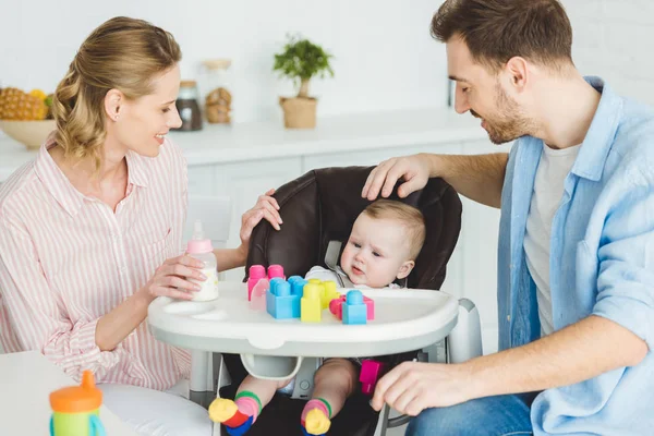 Parents with infant daughter sitting in baby chair with plastic blocks and feeding bottle — Stock Photo