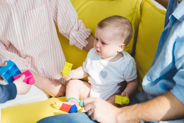 Vista recortada de los padres sentados en el sofá con la hija bebé sosteniendo bloques de plástico - foto de stock