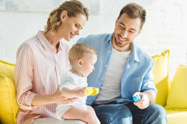 Padres jóvenes sentados en un sofá con una hija pequeña sosteniendo bloques de plástico - foto de stock