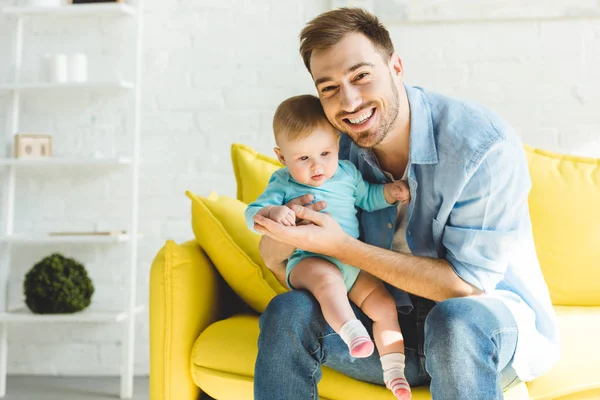 Young smiling father sitting on sofa with infant daughter in hands — Stock Photo