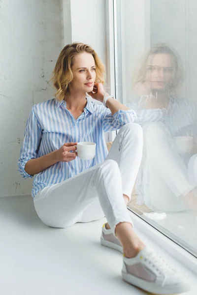 Mujer joven y elegante sentado en el alféizar de la ventana con la taza de café en la mano - foto de stock