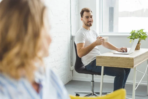 Homme assis à table avec ordinateur portable tout en tenant une tasse de café et en parlant à sa petite amie — Photo de stock