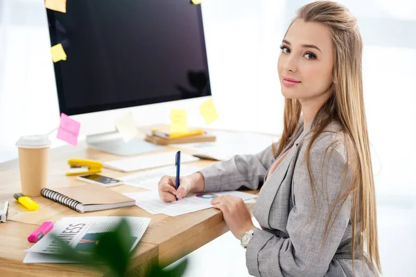 Vista lateral de la mujer gerente de marketing mirando a la cámara mientras hace papeleo en el lugar de trabajo en la oficina - foto de stock