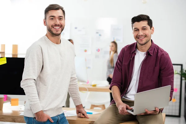 Selective focus of smiling marketing managers with laptop leaning on workplace in office — Stock Photo