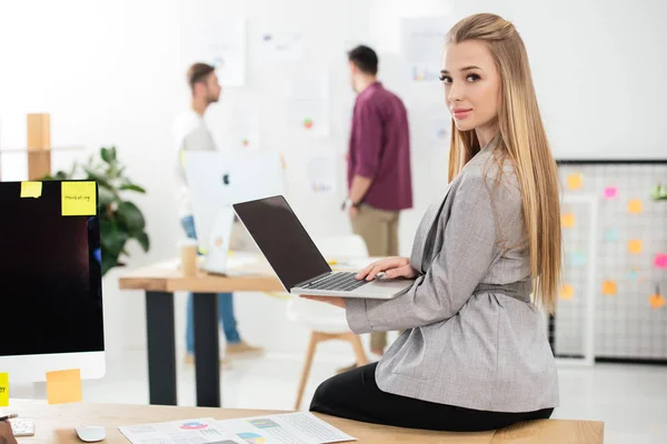 Selektive Fokussierung der jungen Geschäftsfrau mit Laptop auf Kamera im Büro — Stockfoto