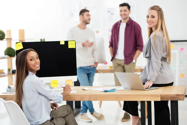 Enfoque selectivo de las empresarias multiétnicas mirando a la cámara en el lugar de trabajo y colegas detrás en la oficina — Stock Photo