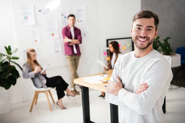 Избирательный фокус улыбающегося менеджера по маркетингу, смотрящего в камеру с коллегами по мультикультурности в офисе — стоковое фото
