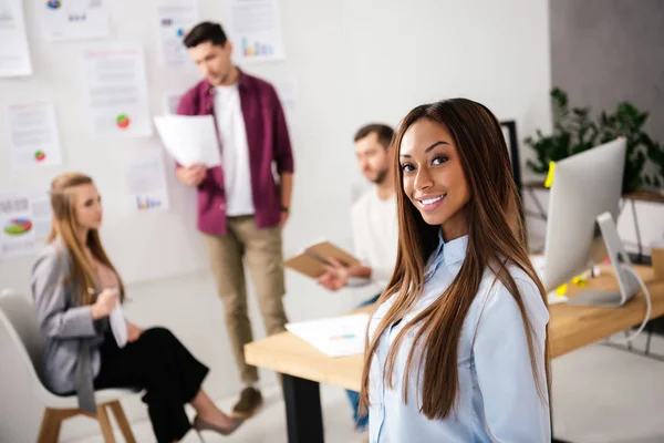 Selektiver Fokus einer lächelnden afrikanisch-amerikanischen Geschäftsfrau, die mit multiethnischen Kollegen im Büro in die Kamera blickt — Stockfoto